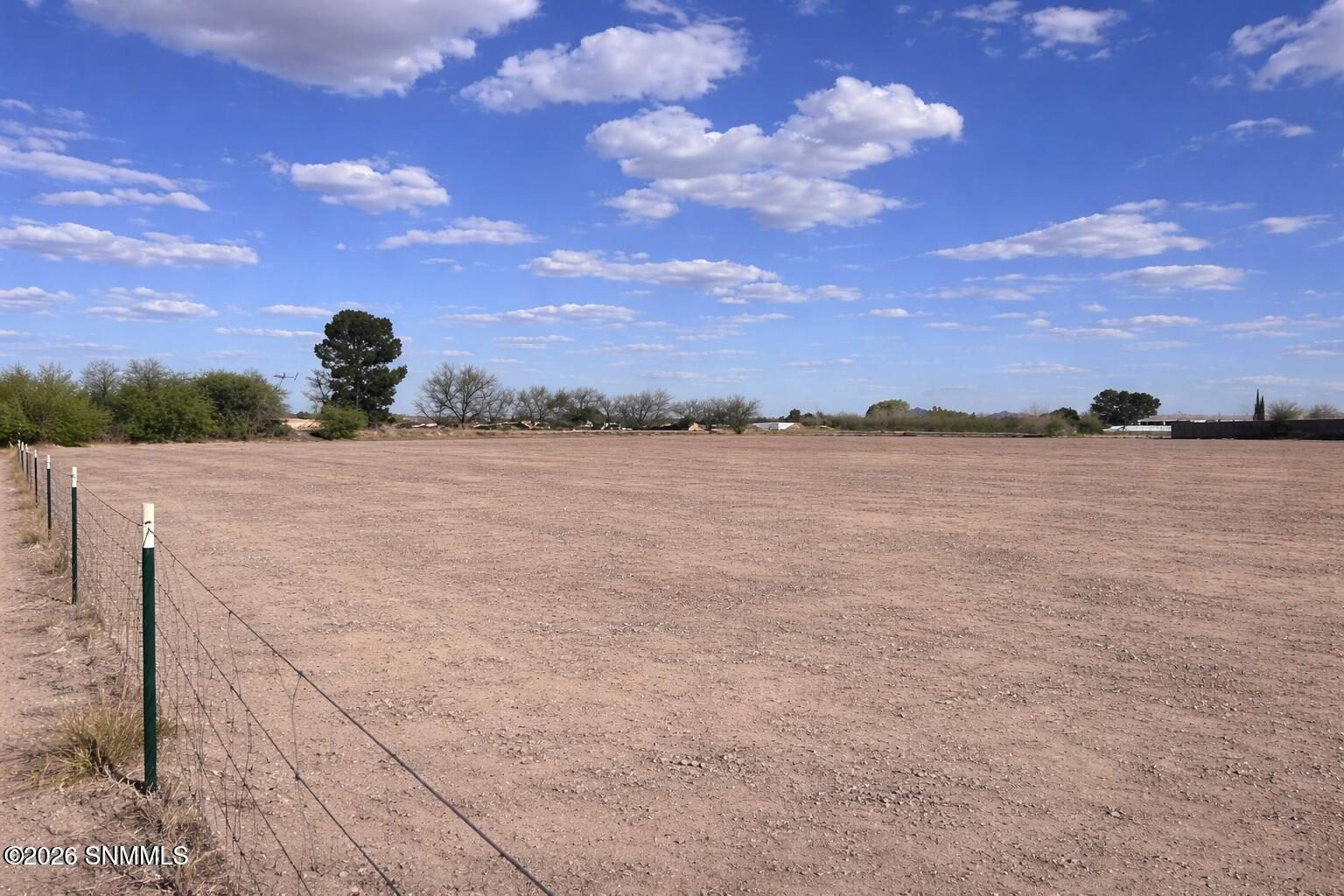 Open dirt lot with wire fencing under a blue sky with scattered clouds and trees along the horizon.