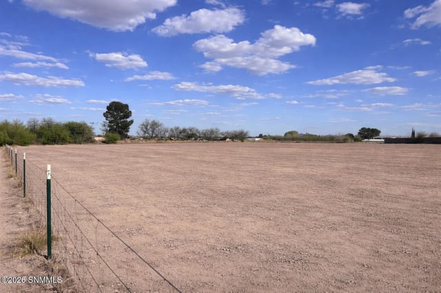 Open dirt lot with wire fencing under a blue sky with scattered clouds and trees along the horizon.
