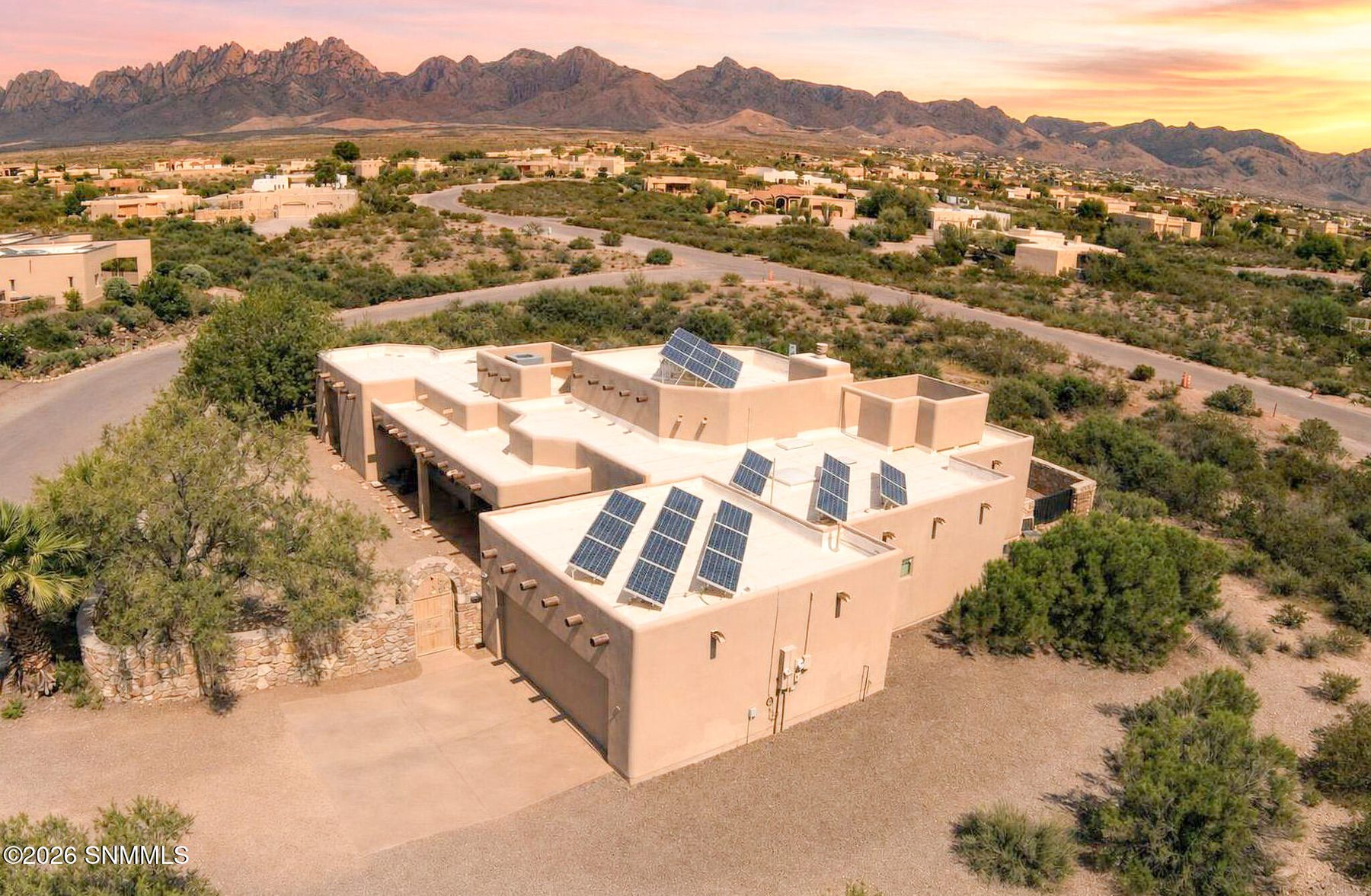 Aerial photo at sunset of a tan stucco Southwestern home with rooftop solar panels, surrounded by desert landscaping, with the Organ Mountains in the background.
