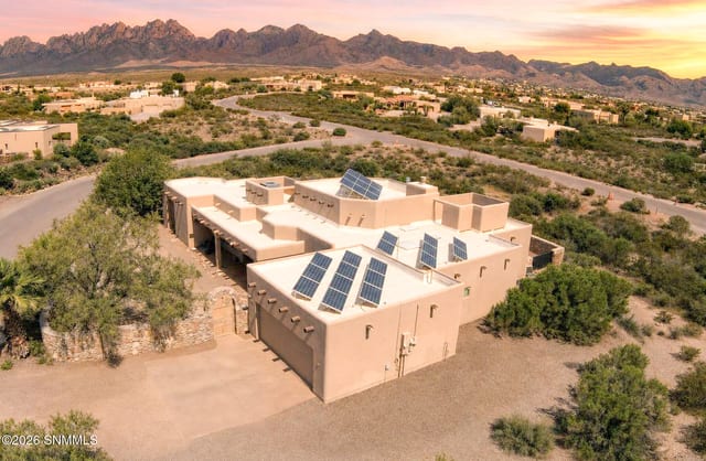 Aerial photo at sunset of a tan stucco Southwestern home with rooftop solar panels, surrounded by desert landscaping, with the Organ Mountains in the background.