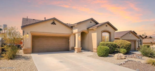 Stucco single-story home with arched entry, two car garage, and desert landscaping at dusk.
