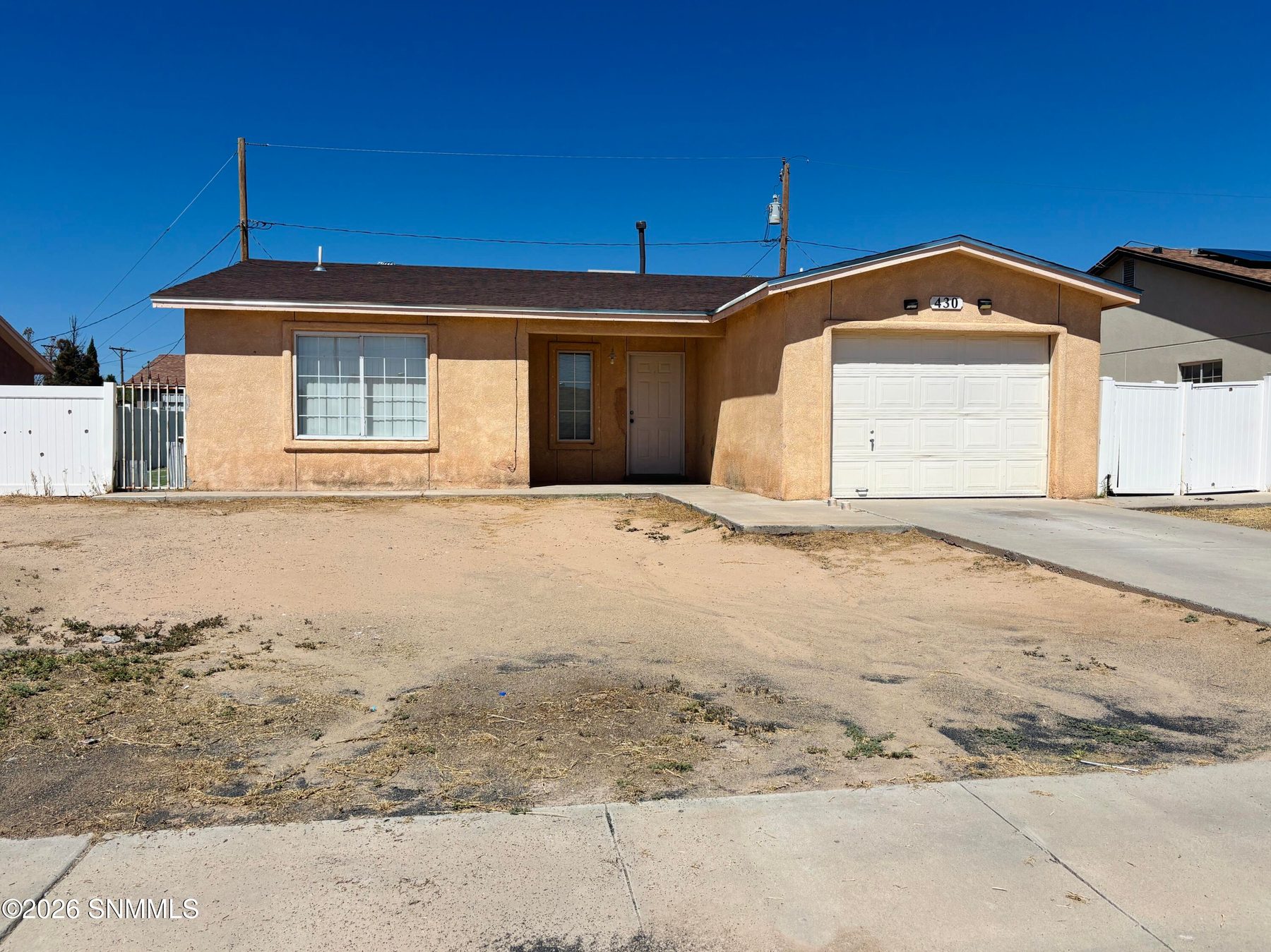 Tan stucco single story home with a white garage door, fenced yard, and dirt front lawn.
