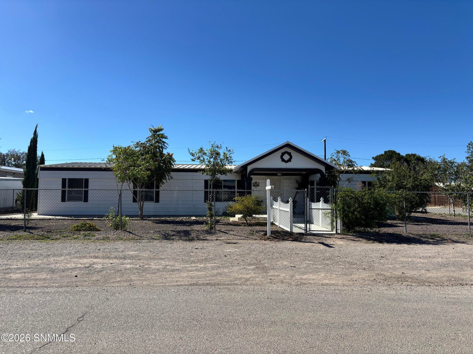 White manufactured home with black trim, metal roof, and a gabled covered entry, enclosed by a chain link and picket fence under a clear blue sky.